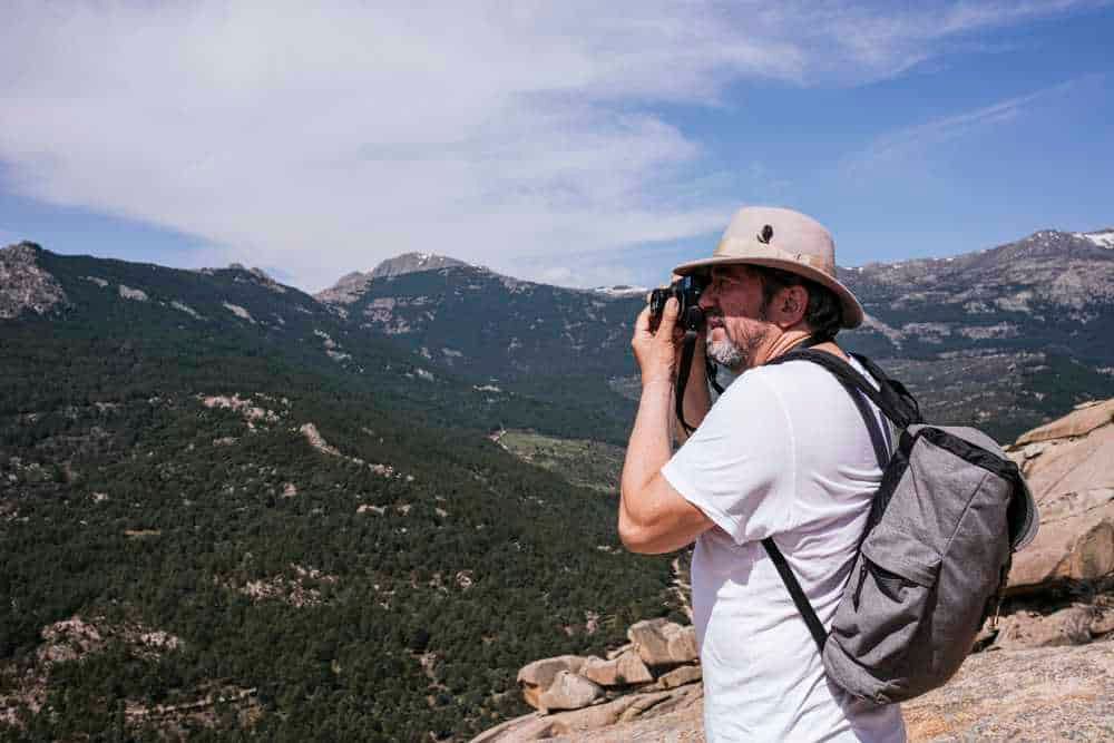 Escursionista che scatta fotografie in montagna con zaino e cappello, esplorando paesaggi naturali e panorami montani.