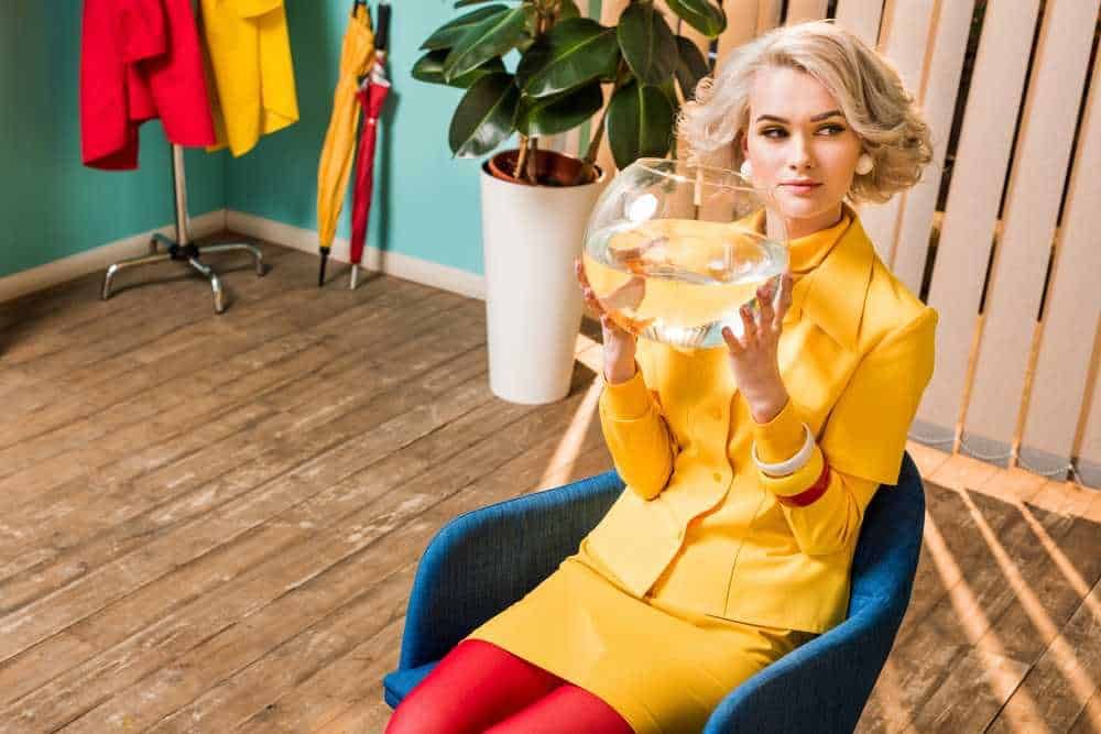 Serena donna in ufficio con abito giallo che tiene un pesce in una ciotola di vetro, ambientazione moderna con piante e arredi colorati, rappresenta innovazione e creatività nel lavoro.