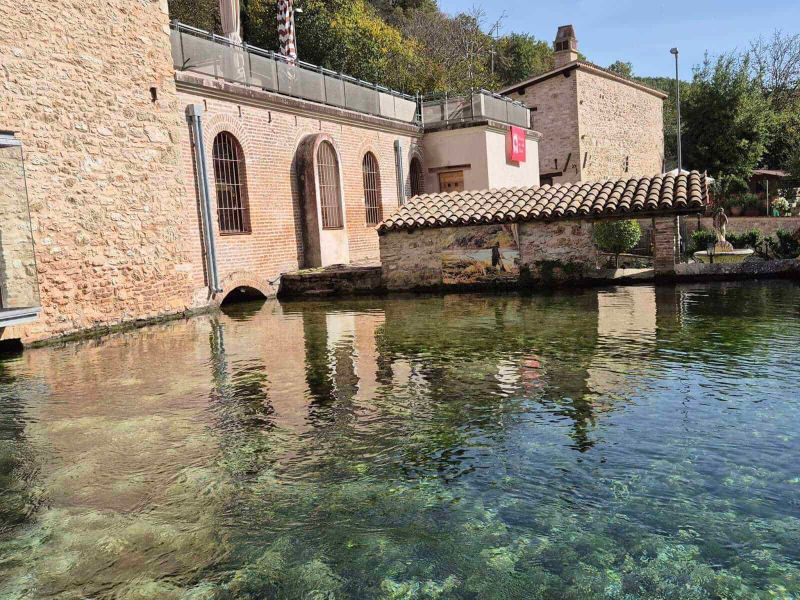 Rasiglia Umbria: Where It Is Located and What to See 2 Rasiglia in umbria: Vecchia centrale idroelettrica ristrutturata con pareti di mattoni e finestre ad arco, situata sul bordo di un lago naturale circondato da vegetazione. Un luogo storico e suggestivo, perfetto per fotografie e visite culturali.