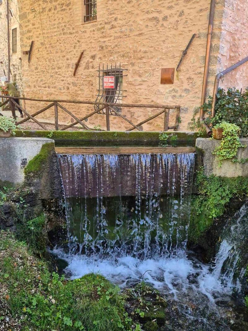 Rasiglia Umbria: Where It Is Located and What to See 5 Arroyo urbano con cascata in pietra lungo un edificio storico in stile toscano, con vivaio di piante e balcone in legno, ambiente rustico e suggestivo per fotografie di paesaggi e natura.