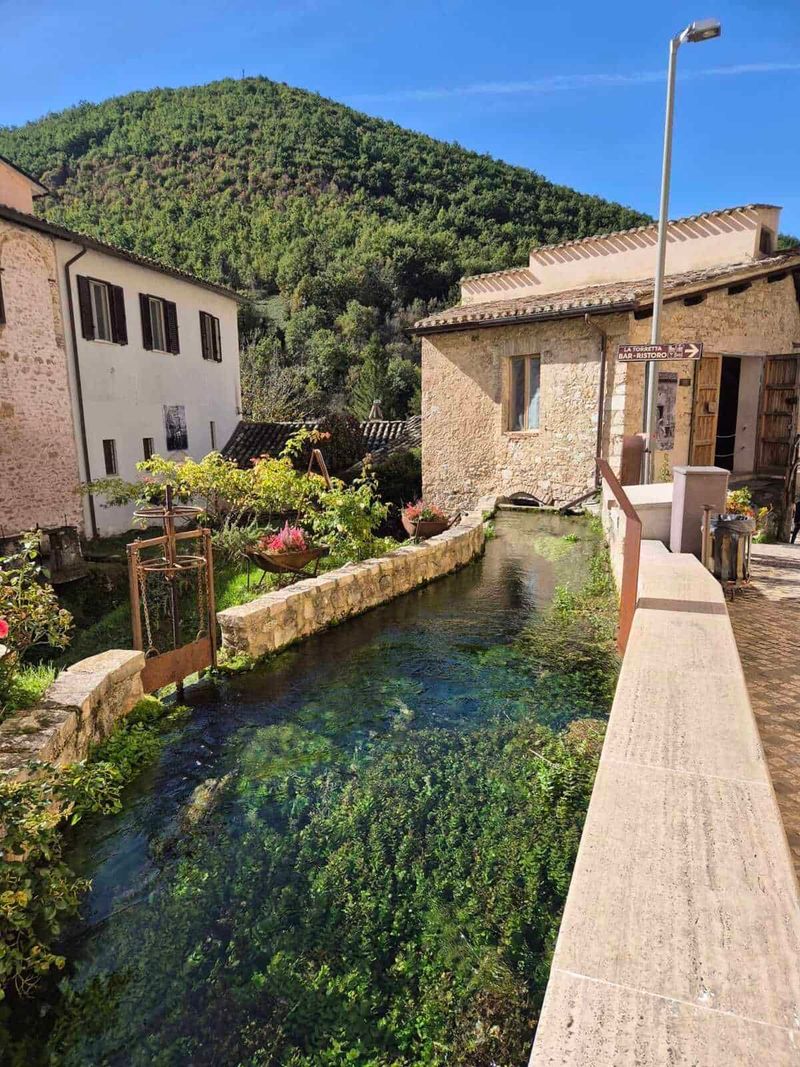 Rasiglia Umbria: Where It Is Located and What to See 4 Fiume in un piccolo villaggio con case in pietra, fiori e una vista sulle colline verdi sotto un cielo azzurro, creando un'atmosfera italiana suggestiva e tranquilla.