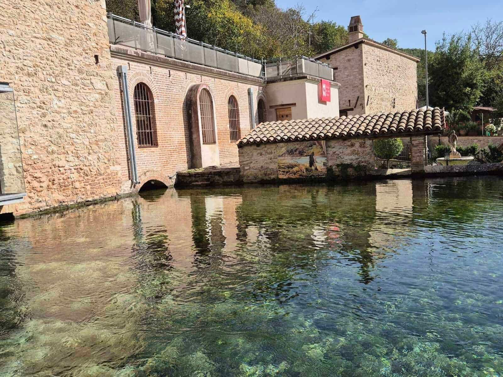 Vecchia centrale idroelettrica ristrutturata con pareti di mattoni e finestre ad arco, situata sul bordo di un lago naturale circondato da vegetazione.