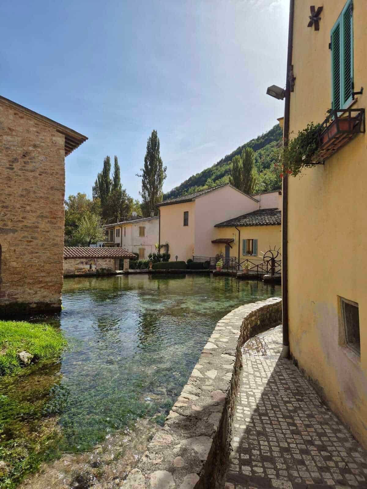 Vecchio paesino italiano con case colorate, mulino e affascinante fontana lungo il fiume, immerso nella natura delle colline, perfetto per relax e fotografia.