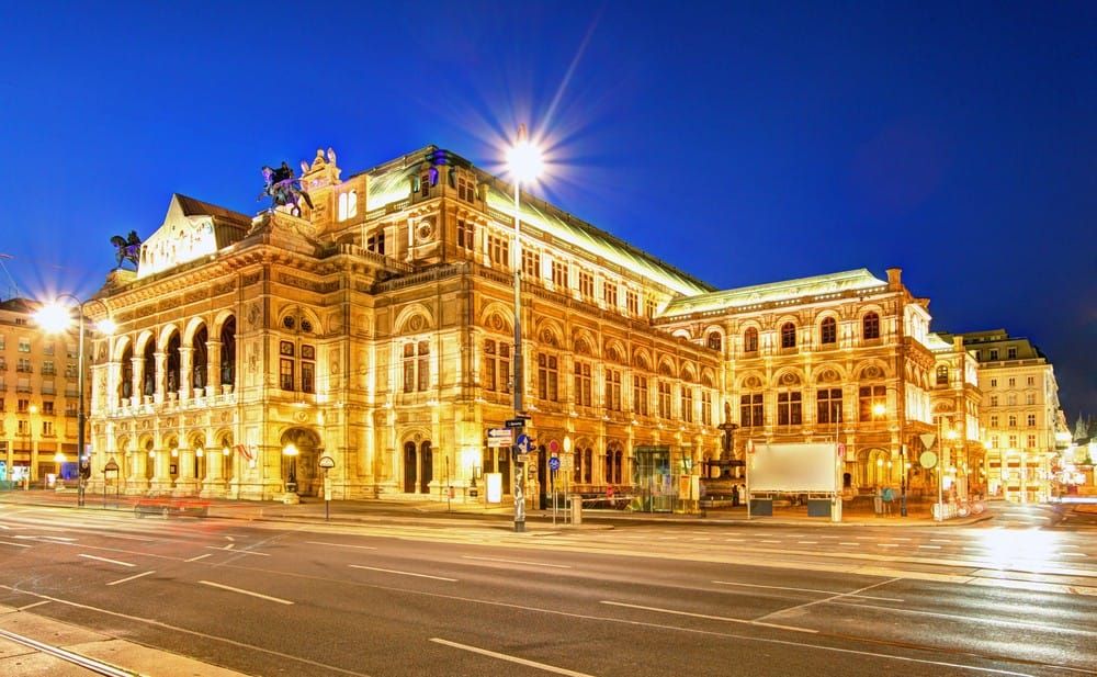 Immagine di un elegante teatro storico di Vienna, illuminato di sera con luci calde, che evidenziano la sua architettura neoclassica e rinascimentale.