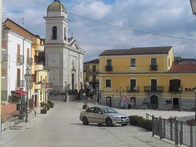 Storica piazza di paese con chiesa barocca, edifici colorati e auto parcheggiate, scena tranquilla di una città italiana. Scene di vita quotidiana e architettura tradizionale in un centro storico.