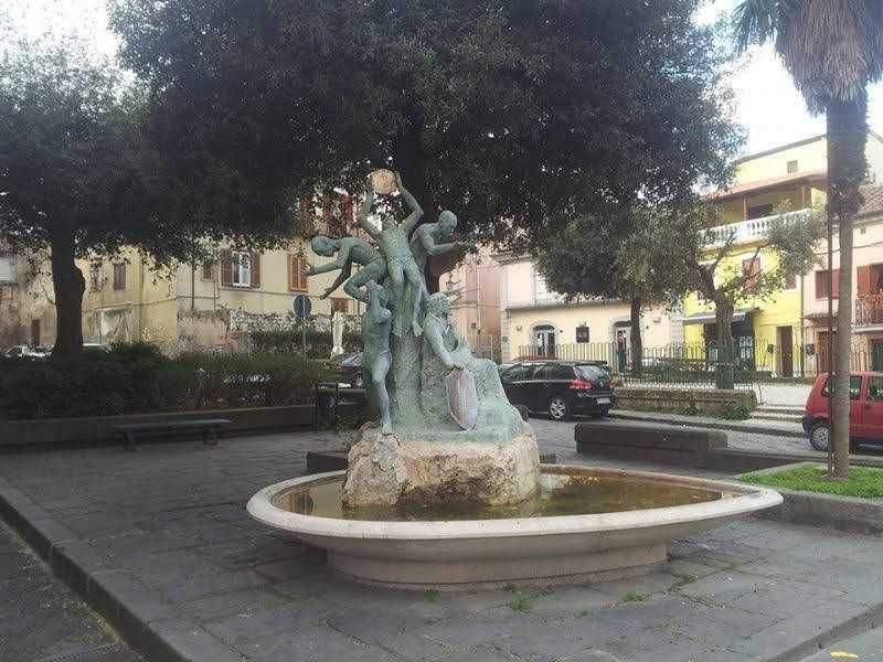 Luoghi di padre pio: Fontana decorativa nel centro città con sculture di figure umane in bronzo, circondata da un bacino d'acqua, con edifici storici e alberi sullo sfondo, un esempio di arte pubblica italiana.