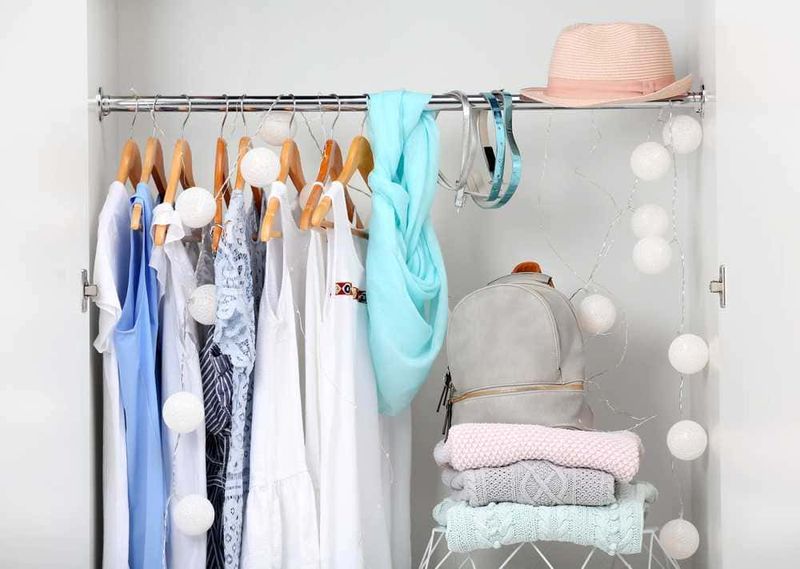 Vestiti appesi o piegati nell'armadio? 5 Vestiti estivi bianchi e azzurri appesi in armadio con decorazioni di luci a sfere. Accessori come cappelli e zaini sono visibili su una mensola per un guardaroba ordinato e stiloso.