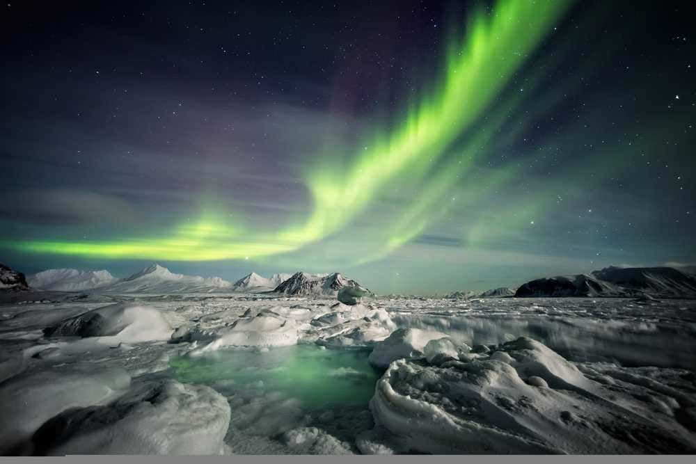 Artico e antartide: Aurora boreale sopra paesaggio ghiacciato artico invernale, montagne innevate e cielo stellato, spettacolo naturale nordico, atmosfere magiche, fotografia paesaggistica, fenomeno spettacoloso, turismo avventuroso, natura selvaggia.