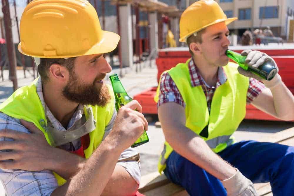 Becceri da cantiere con caschi di sicurezza e giubbotti gialli bevendo birra durante la pausa lavoro all'aperto in un sito di costruzione.
