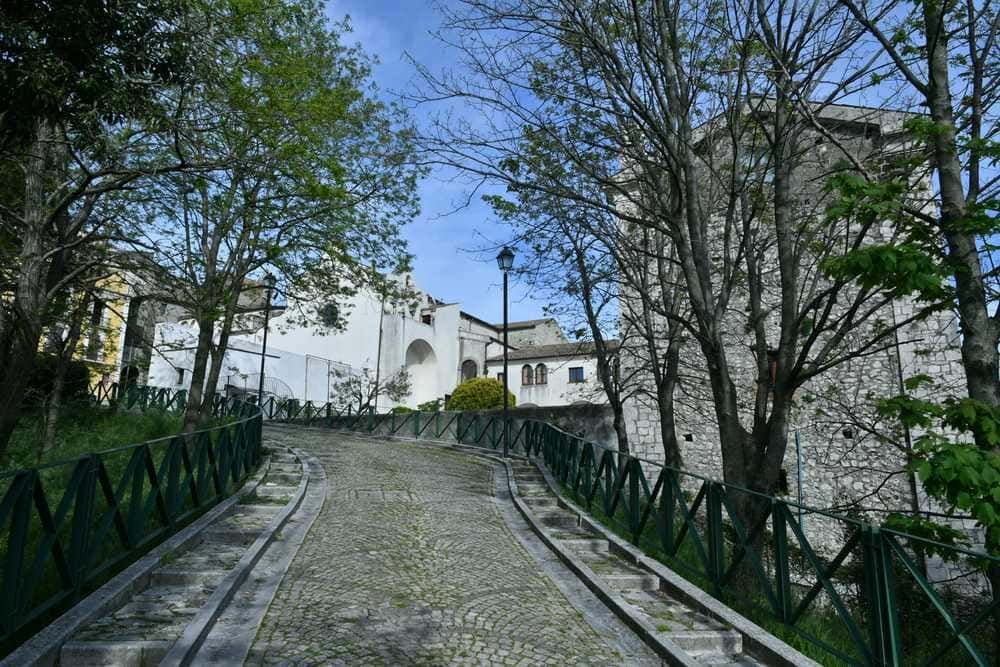 Pendenza di strada in pietra con alberi verdi e edifici storici in stile medievale in background, sotto un cielo azzurro, zona turistica e storica di un borgo italiano, ideale per appassionati di storia e natura.