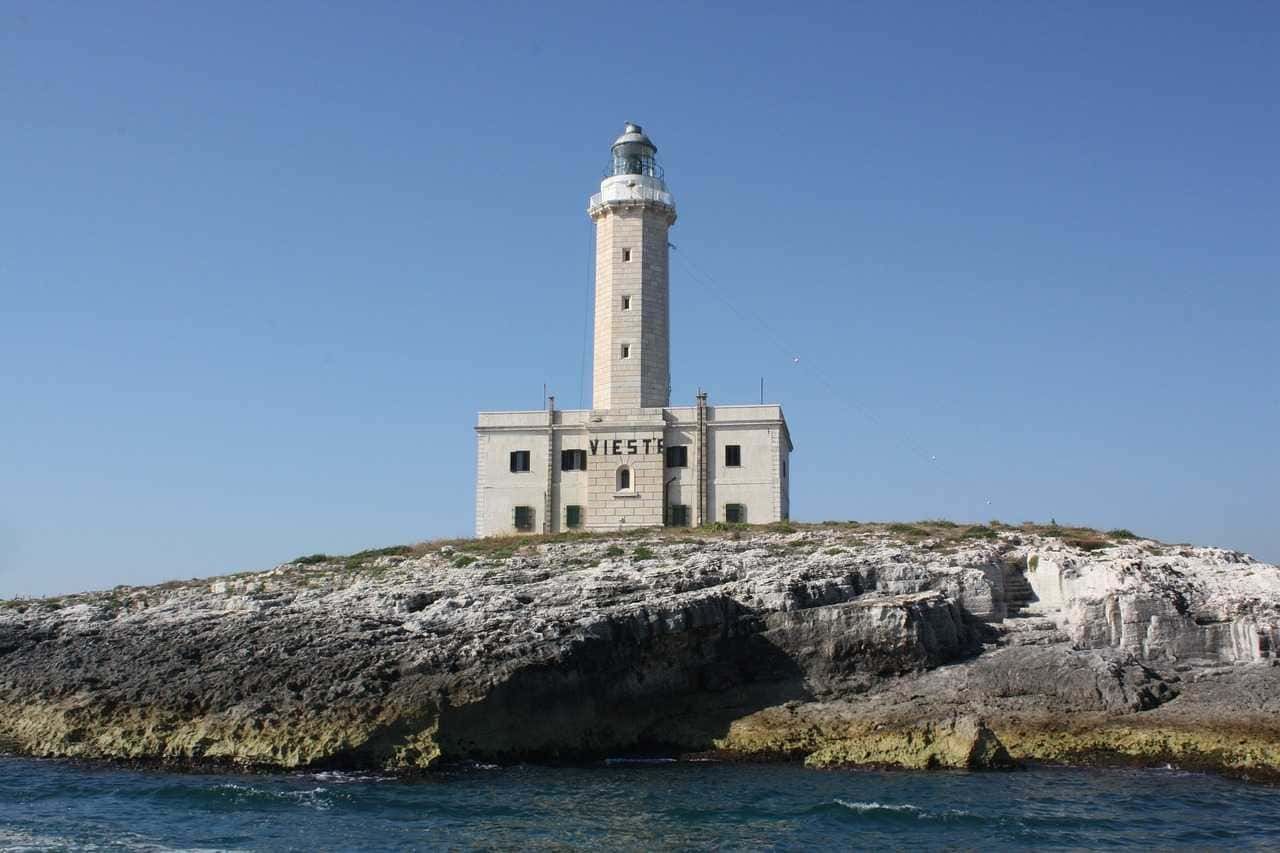 Faro di Vieste situato sulla scogliera a picco sul mare, simbolo del promontorio di Vieste, in Puglia, Italia, con cielo azzurro e acque cristalline.