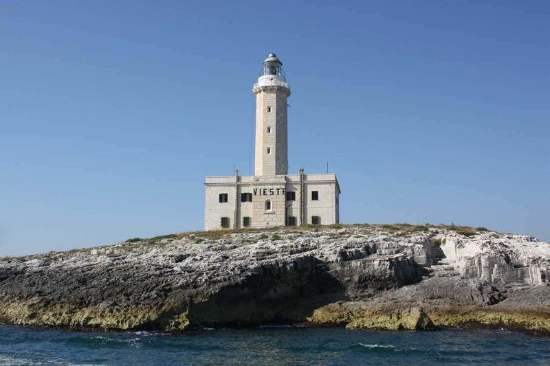 È il momento giusto per prenotare le tue vacanze estive 3 Faro di Vieste situato sulla scogliera a picco sul mare, simbolo del promontorio di Vieste, in Puglia, Italia, con cielo azzurro e acque cristalline.