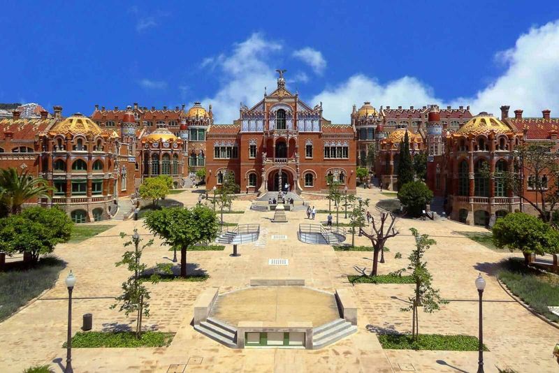 Ospedale Sant Pau Barcellona da visitare 2 Ospedale di sant pau: Castello in stile modernista, edificio storico affascinante con dettagli architettonici elaborati, situato in un parco verde con alberi e sentieri, sotto un cielo azzurro raggiante.