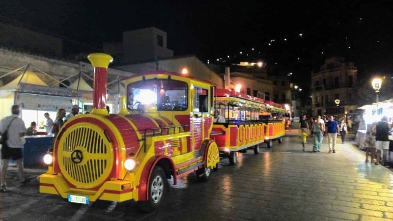 Castellammare del Golfo attrazioni e dintorni 4 Colorato trenino turistico a trazione elettrica per città, perfetto per visite guidate e gite notturne, circondato da persone e negozi vivaci, in una piazza affollata e illuminata.