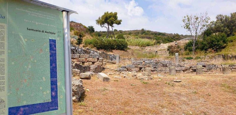 Scavi di Velia Paestum cosa vedere in 2 ore 6 Resti archeologici dell'antico santuario di Asclepio in un sito immerso nella natura, con pannello informativo e paesaggio collinare circostante. Ideale per appassionati di archeologia e storia antica.