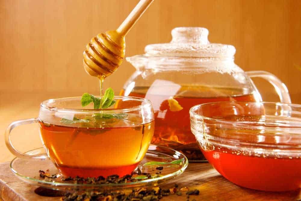 Miele e tisana floreale in tazza di vetro con teiera di vetro sul tavolo in legno, concept di infusi naturali e benessere.