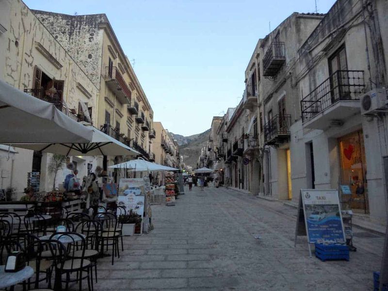 Castellammare del Golfo attrazioni e dintorni 3 Vicolo pittoresco con caffè all'aperto e negozi in un borgo storico italiano con edifici antichi e montagne sullo sfondo.