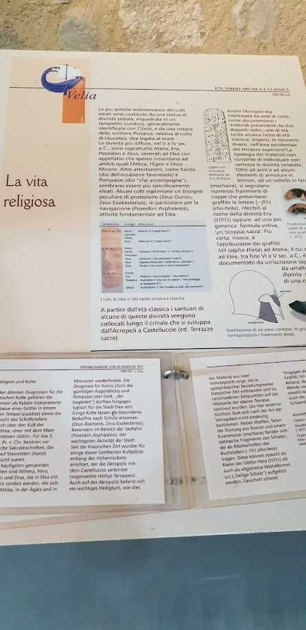 Scritto un testo religioso su un quaderno aperto con immagini di simboli religiosi e testo in italiano e tedesco, in un ambiente con pareti di pietra.