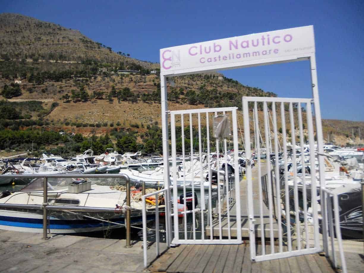 Cn Club Nautico Castellammare di Stabia, porto turistico con barche e yacht, ingresso con cancellata bianca, paesaggio montano sullo sfondo, cielo sereno, immagine di arredo marittimo, vacanze e nautica.