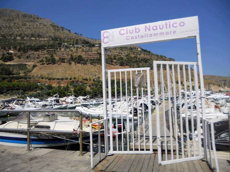 Castellammare del Golfo attrazioni e dintorni 5 Cn Club Nautico Castellammare di Stabia, porto turistico con barche e yacht, ingresso con cancellata bianca, paesaggio montano sullo sfondo, cielo sereno, immagine di arredo marittimo, vacanze e nautica.