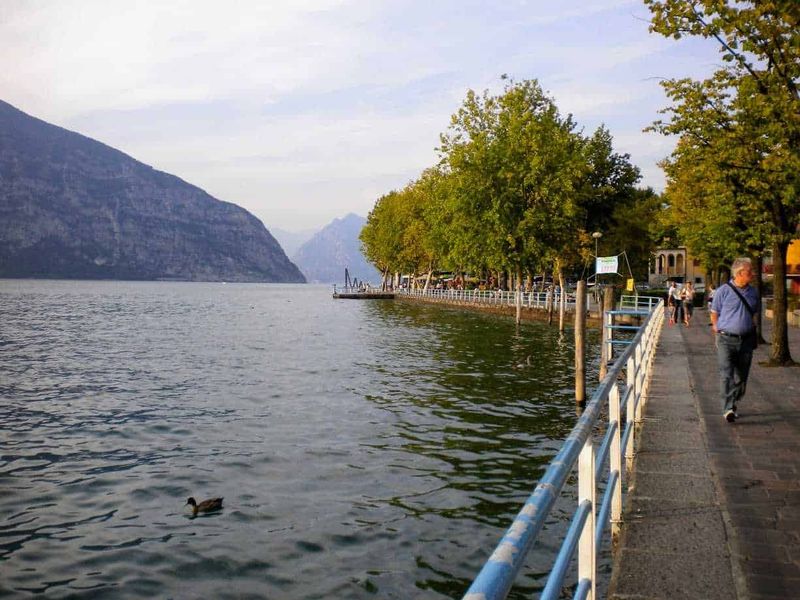 Cosa Vedere a Iseo città sul Lago di Iseo: centro storico, castello e lungolago 3 Lungolago con passeggiata in riva al lago, alberi e montagne sullo sfondo, persone che passeggiano e anatroccoli che nuotano in acqua, scenario tranquillo e pittoresco, ideale per fotografia e relax.