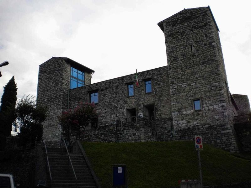 Cosa Vedere a Iseo città sul Lago di Iseo: centro storico, castello e lungolago 4 Castello Oldofredi, medievale in pietra con torri, situato su una collina, con bandiera italiana, palazzi storici, vista panoramica, in Italia, esempio di architettura storica e patrimonio culturale.