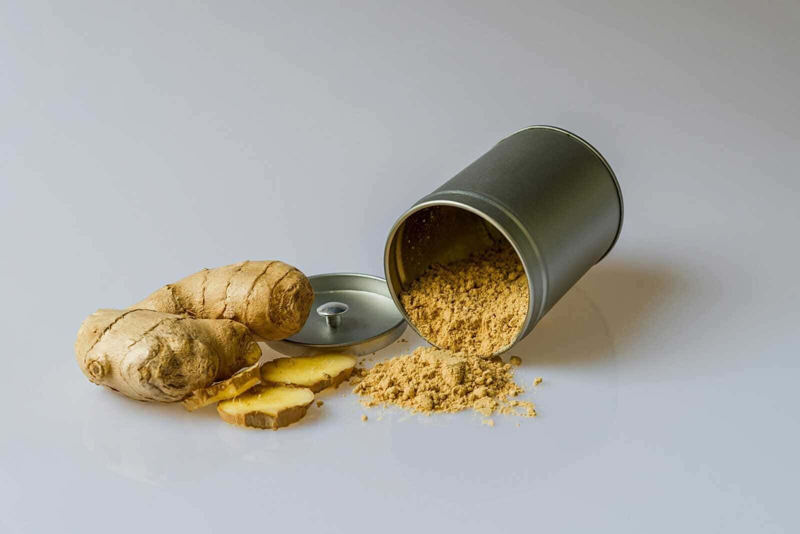 Polvere di zenzero in scatola con radici fresche di zenzero e alcune fette di zenzero fresco disposte su uno sfondo chiaro.