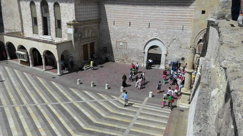 Assisi cosa vedere: itinerario tra arte e patrimonio UNESCO (2026) 6 Piazza storica di Assisi con persone, architettura antica e pavimentazione decorativa, scena urbana nel centro di una città italiana