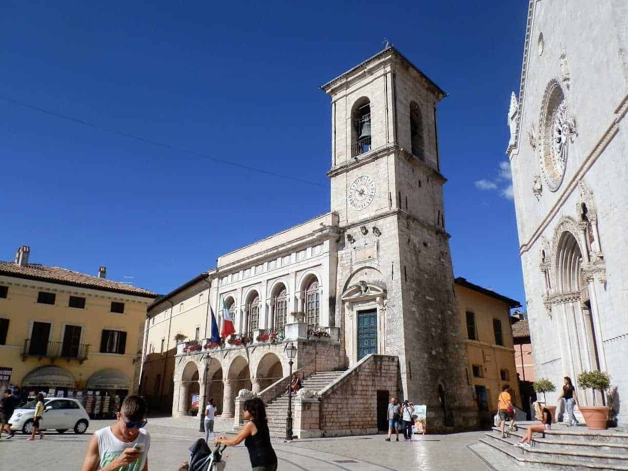 Campanile storico in piazza con persone e auto, edifici antichi con facciata decorata, cielo blu e giornata di sole, atmosfera vivace nel centro di un borgo italiano.