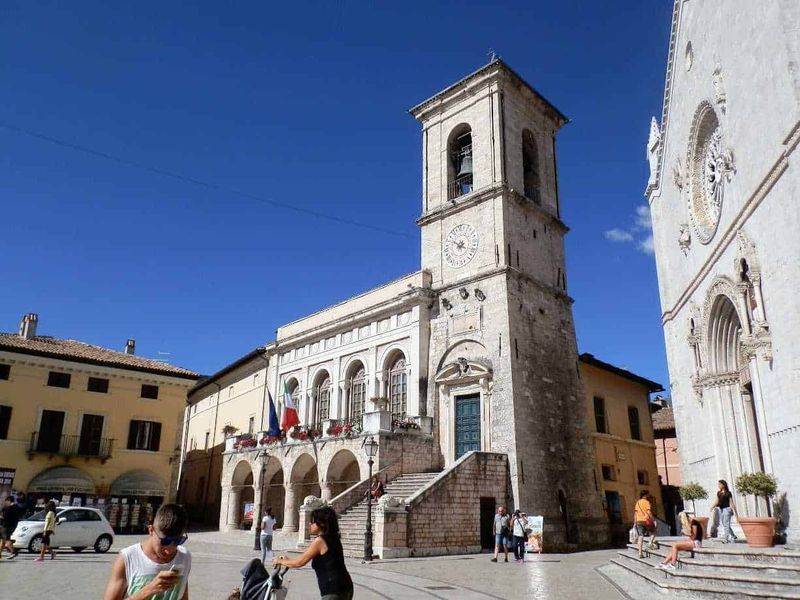 6 destinazioni da sogno in Italia da scoprire 6 Campanile storico in piazza con persone e auto, edifici antichi con facciata decorata, cielo blu e giornata di sole, atmosfera vivace nel centro di un borgo italiano.