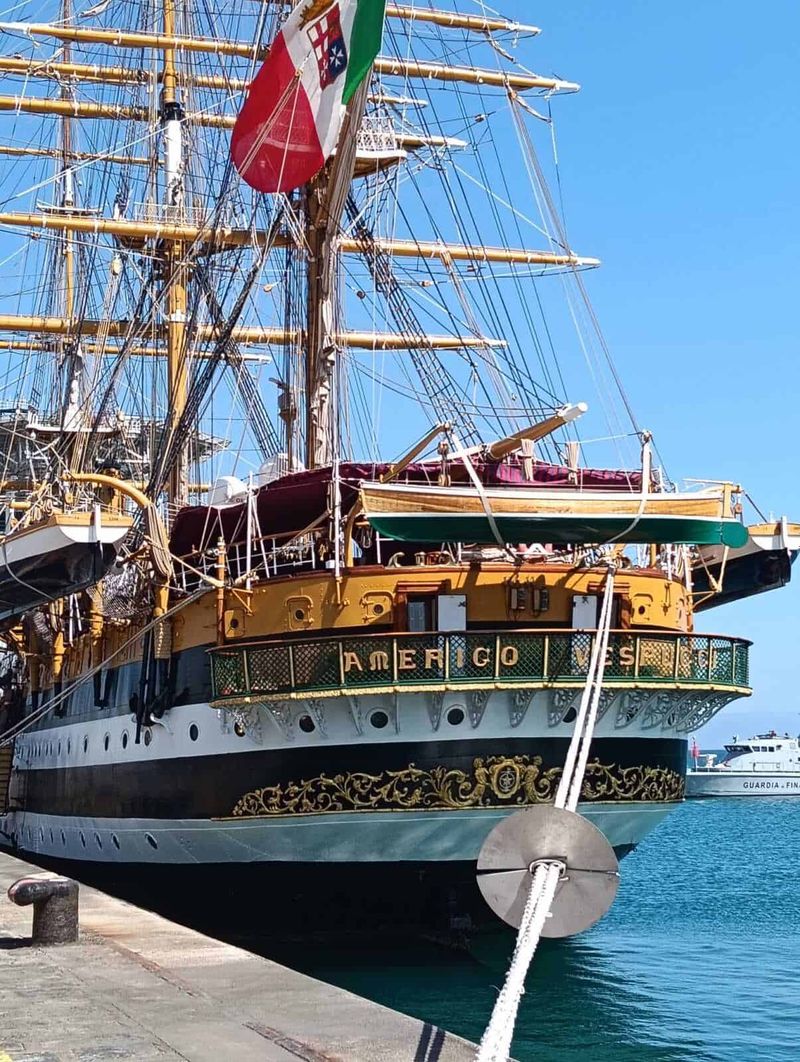 Amerigo Vespucci a Napoli 2025: cosa sapere 3 Antico veliero con vele spiegate ormeggiato al porto, bandiera italiana visibile, imbarcazione storica e di pregio, cielo azzurro e acqua blu sullo sfondo.