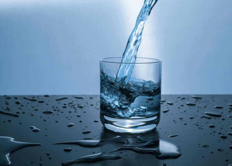 Acqua pura, salute sicura: perché scegliere un depuratore d'acqua per la tua casa 4 Acqua che viene versata in un bicchiere trasparente con gocce e schizzi sparse sulla superficie nera, evidenziando purezza e freschezza.
