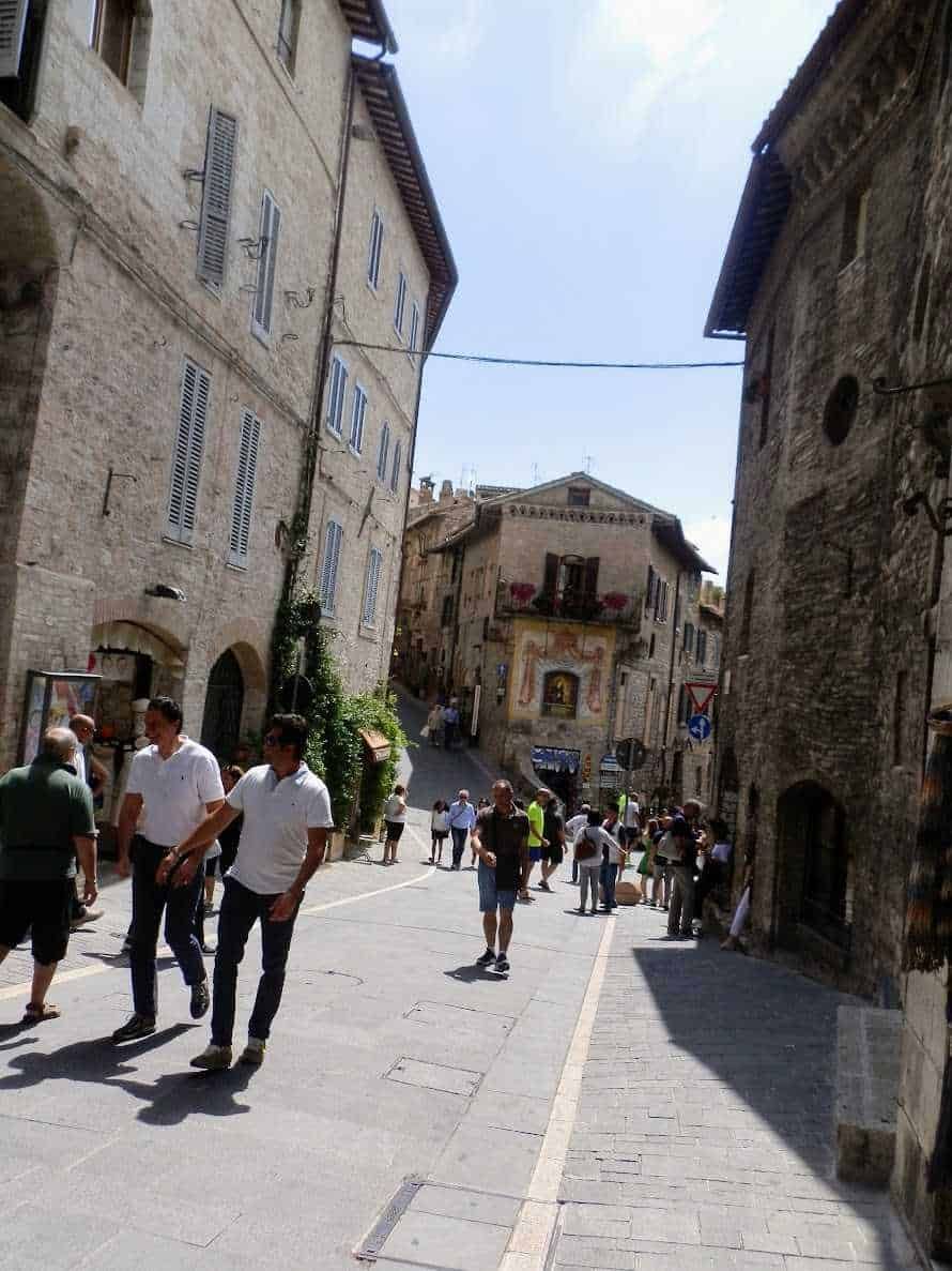 Vicolo in centro storico di Assisi con persone che passeggiano, edifici in pietra e decorazioni murali, atmosfera mediterranea, strada pedonale, architettura tipica italiana.