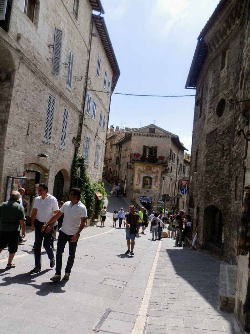 Assisi cosa vedere: itinerario tra arte e patrimonio UNESCO (2026) 2 Vicolo in centro storico di Assisi con persone che passeggiano, edifici in pietra e decorazioni murali, atmosfera mediterranea, strada pedonale, architettura tipica italiana.