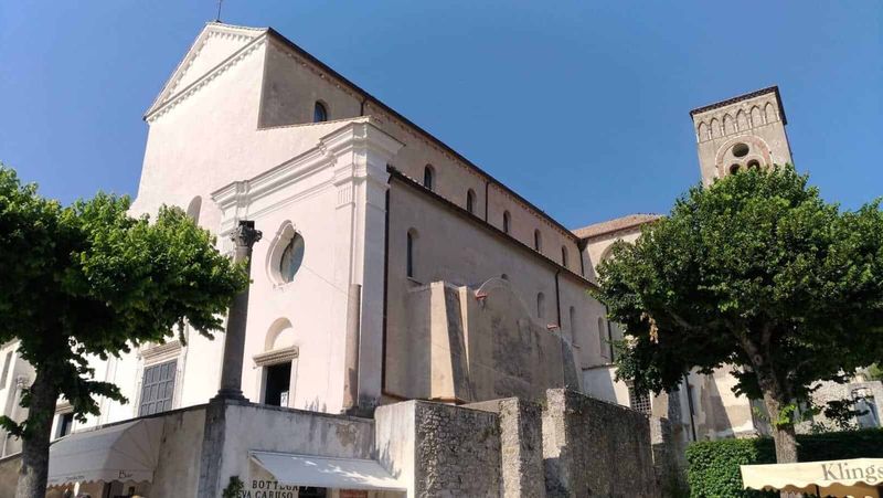 Stili dei portoni a Ravello: dal Medioevo al rustico 3 Antico edificio religioso con torri e facciata bianca, circondato da alberi verdi e situato in un centro storico soleggiato.