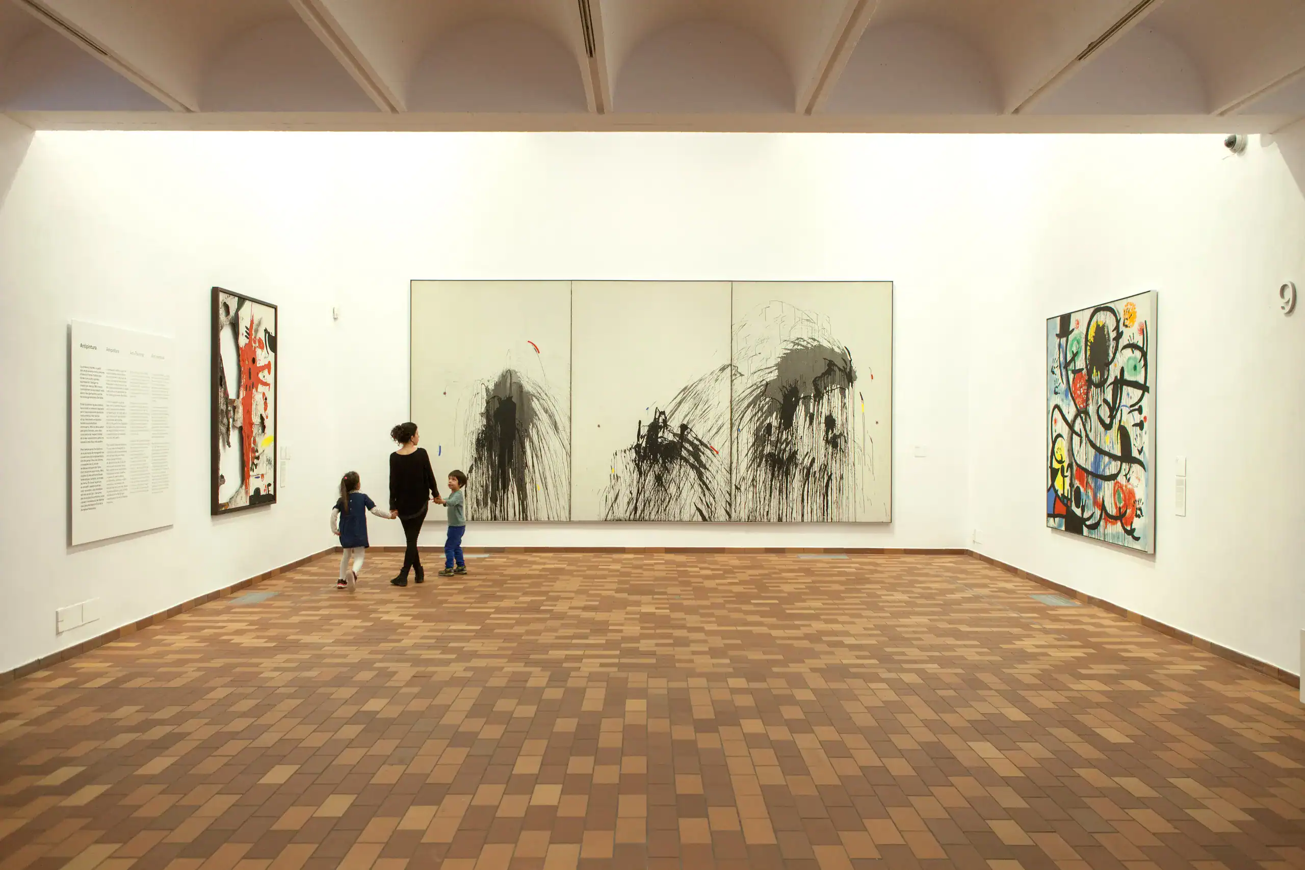 Montjuïc Barcellona cosa vedere in 1 giorno 7 Dipinto astratto bianco e nero in una galleria d'arte, con due bambini e un adulto che osservano l'opera, pavimento in piastrelle di mattone, pareti bianche e illuminazione naturale.