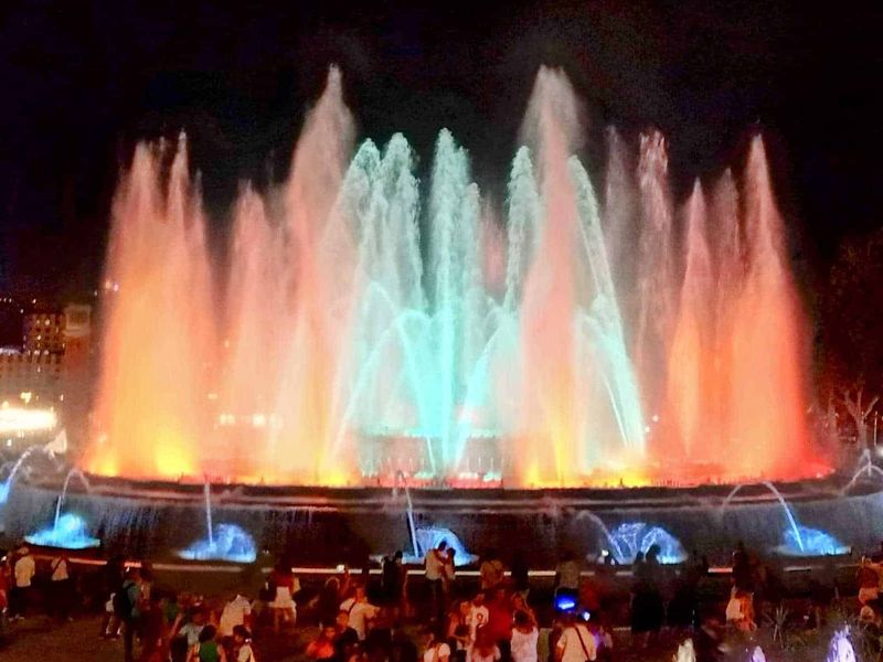 Montjuïc Barcellona cosa vedere in 1 giorno 3 Faro luminoso con giochi d'acqua e luci colorate, spettacolo serale in una piazza, atmosfera festiva, fontane danzanti illuminate, momento di intrattenimento per famiglie e turisti.