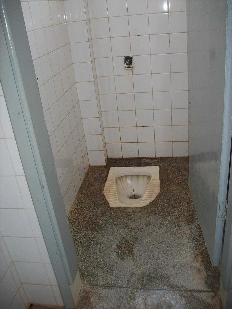 Toilette turca o europea? Differenze, benefici per la salute e quale scegliere 3 Scarico pertoilettes tradizionale in bagno pubblico con piastrelle bianche e pavimento in cemento, senza sedile. Ambiente usato o in fase di ristrutturazione.