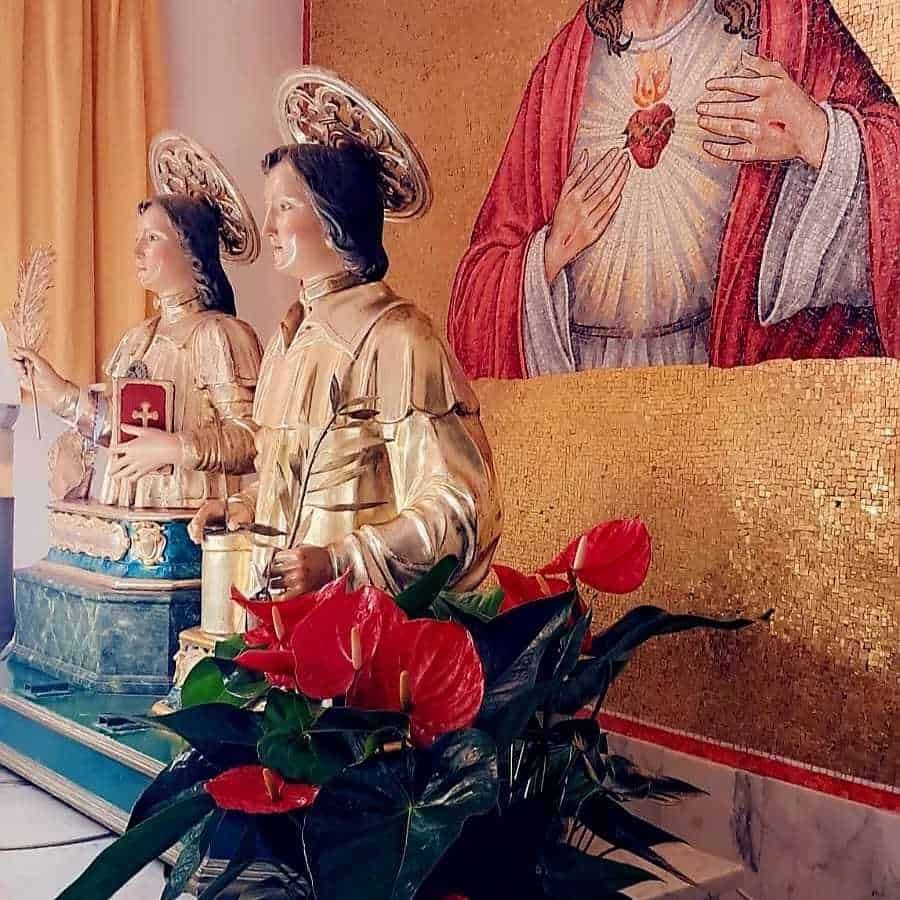 Statuette religiose raffiguranti santi con dettagli dorati, decorazioni natalizie con piante verdi e fiori rossi, in ambiente sacro con mosaici dorati e iconografia religiosa.