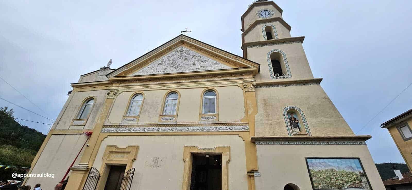 Antico municipio di montagna con scuola cattolica, architettura storica e campanile alto in pietra, dettagli decorativi e affreschi esterni, situato in un pittoresco paesaggio collinare.