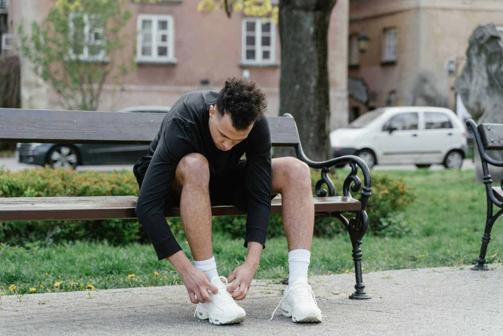 Scarico e indossando scarpe da ginnastica su una panchina nel parco in città. Uomo sportivo si prepara per l'allenamento all'aperto, circondato da alberi e edifici residenziali.
