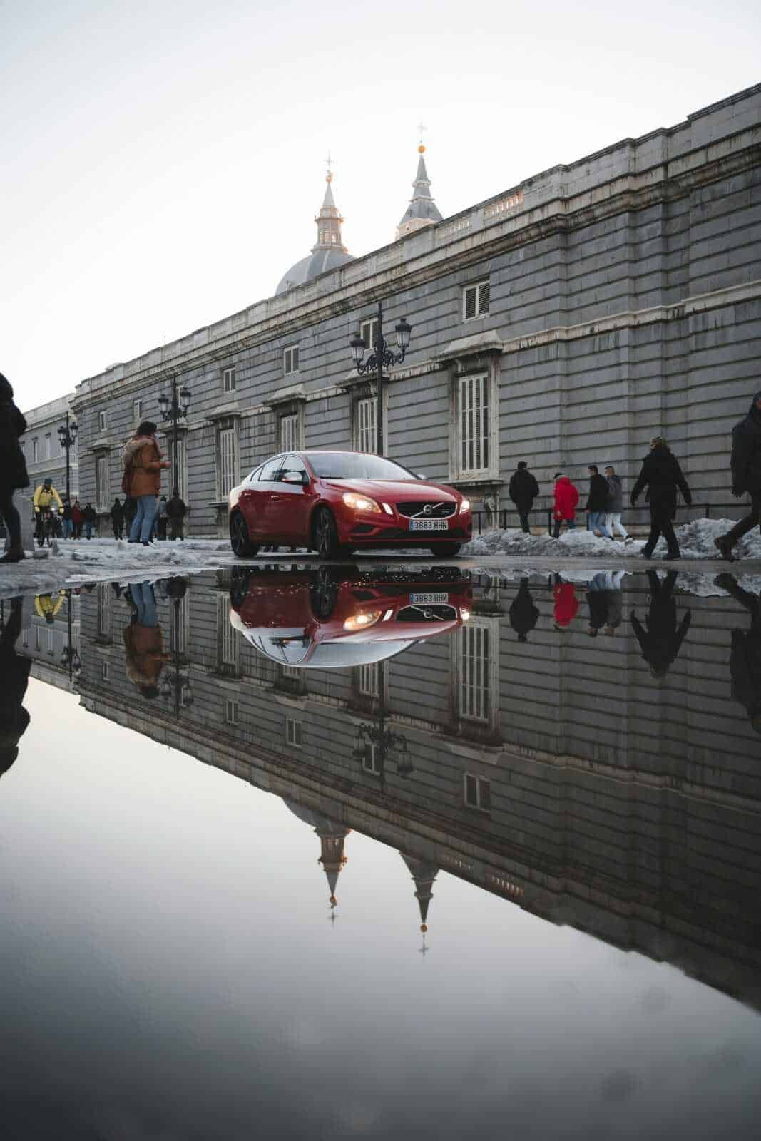Auto rossa parcheggiata vicino a un edificio storico con riflesso sull'acqua, scena urbana con passanti, architettura antica e strade trafficate, in un contesto di città europea.