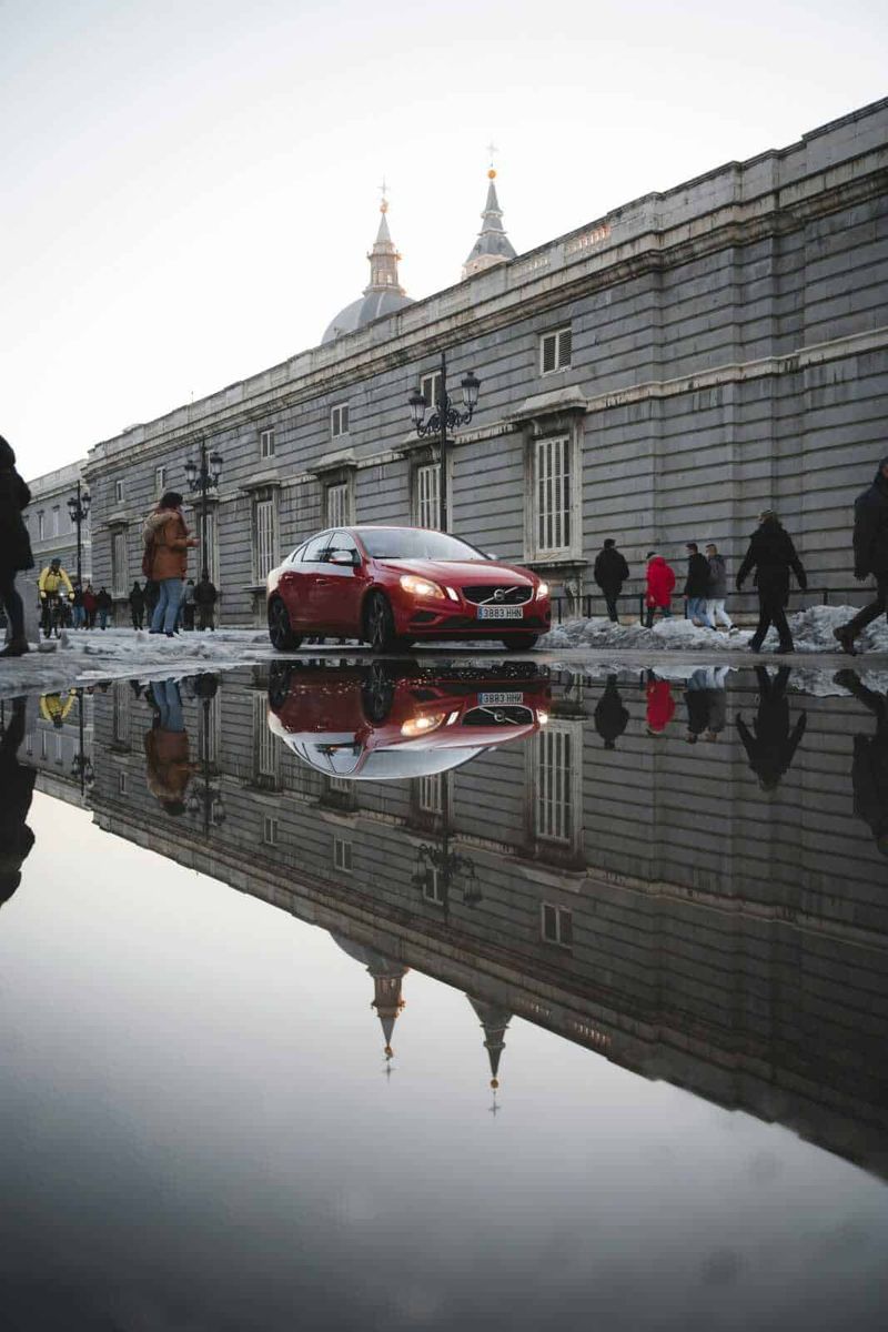 Come guidare correttamente nelle aree allagate 2 Auto rossa parcheggiata vicino a un edificio storico con riflesso sull'acqua, scena urbana con passanti, architettura antica e strade trafficate, in un contesto di città europea.