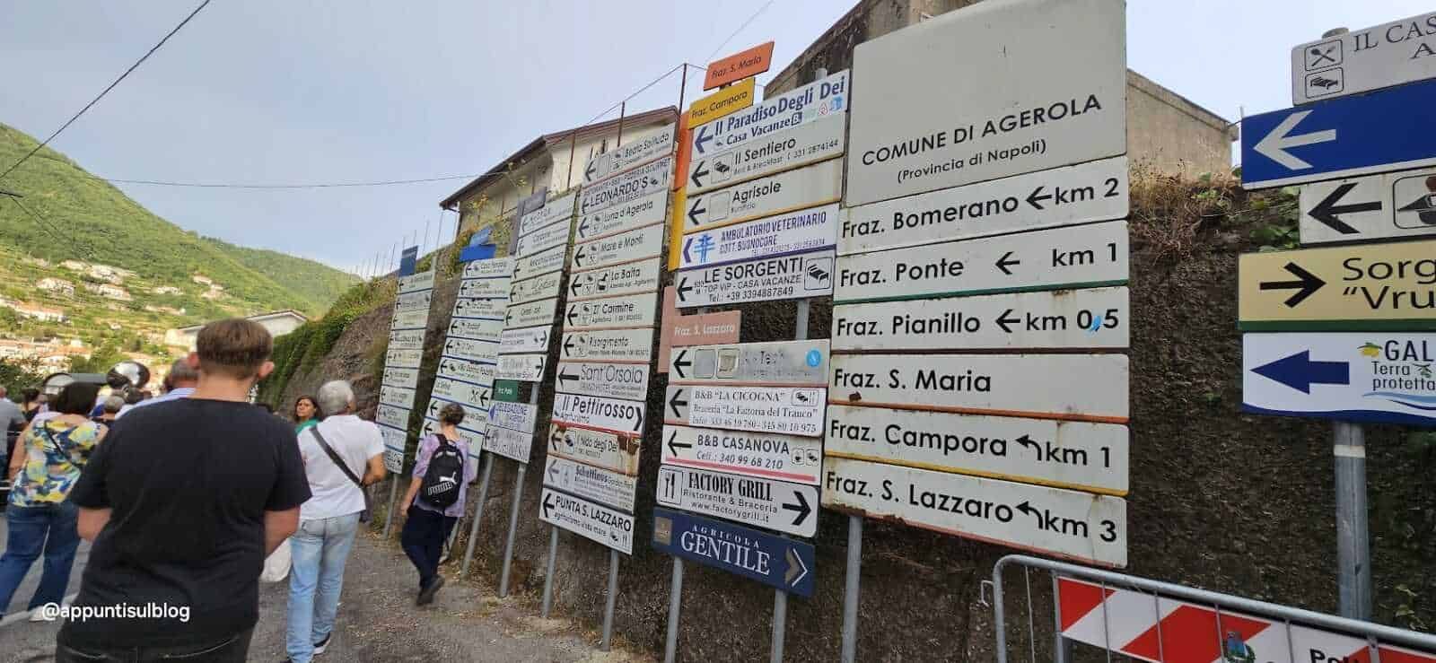 Segnali stradali con indicazioni di località e frazioni a Agerola, Italia, in un paesaggio montano. Persone che camminano lungo una strada di montagna; cartelli turistici e di indicazioni stradali.
