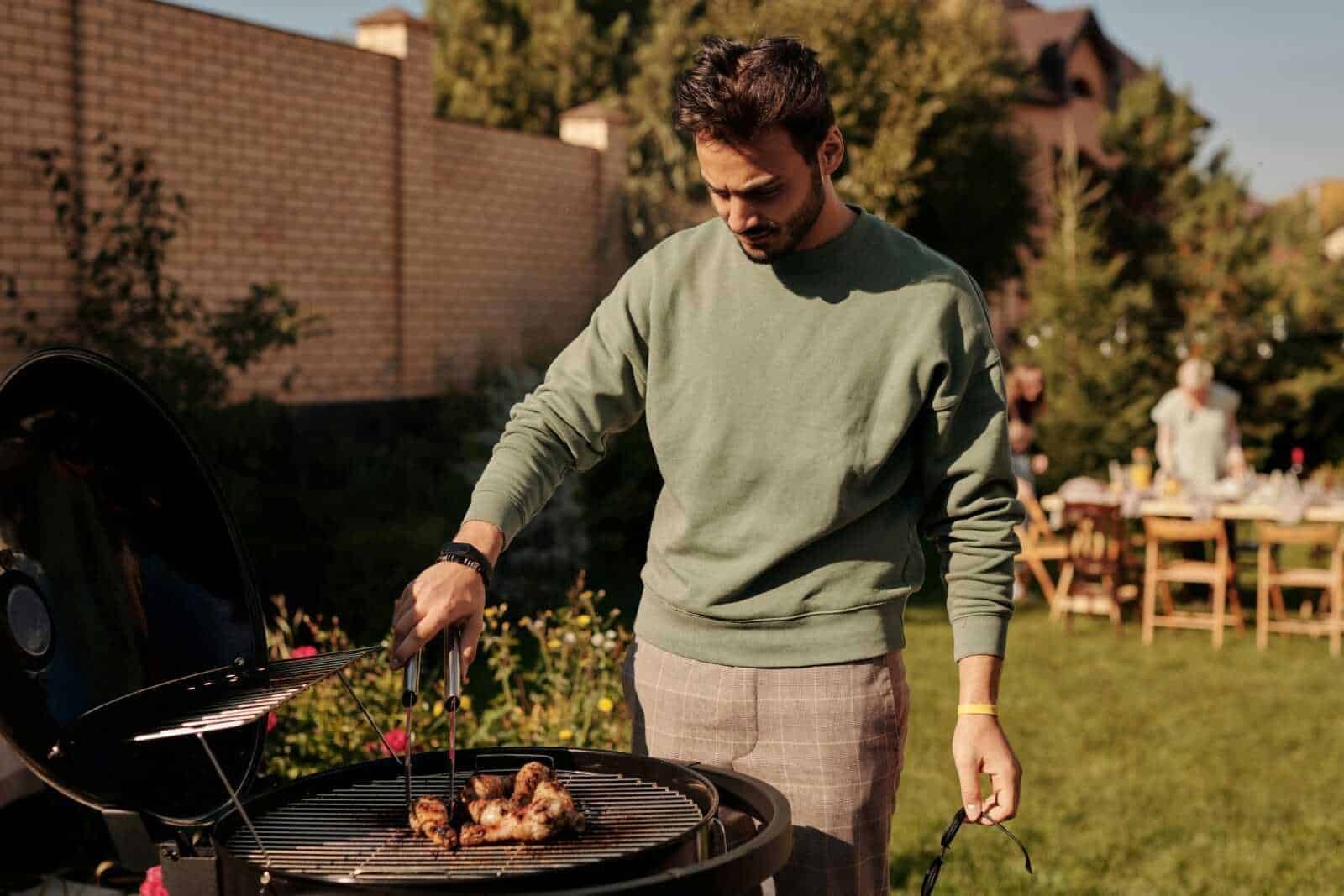 Ferragosto in italia: Grigliata all'aperto in giardino durante un barbecue estivo, con un uomo che controlla le carne sulla griglia, atmosfera conviviale e momenti di relax all'aperto.
