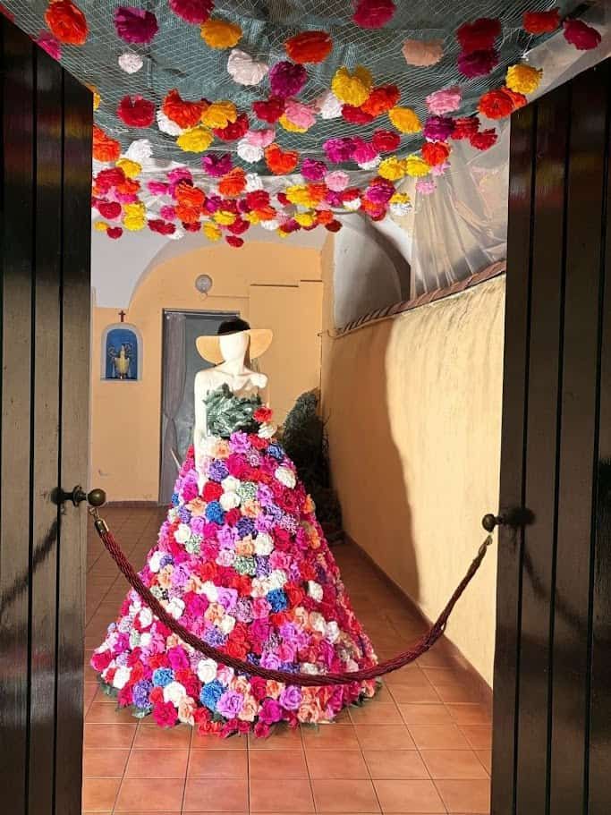 Festa dei fiori Monte Isola 2025 dopo 10 anni 8 Colorata installazione floreale ispirata alle spose di lusso, decorata con un ampio vestito di fiori e un cappello di paglia, circondata da un soffitto di fiori colorati in un ambiente accogliente.