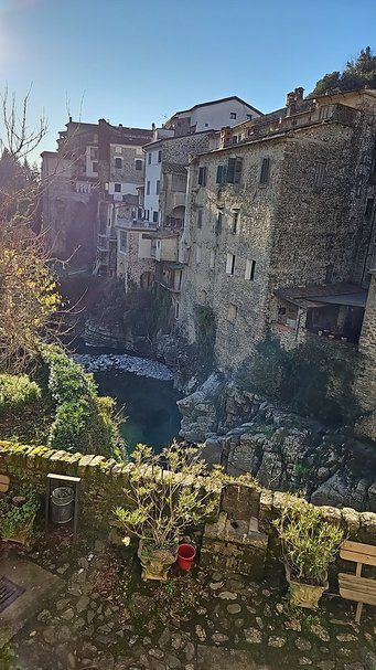 Antica domu appoggiata su scogliera con vista sul fiume, case in pietra e terrazze con piante, ambientazione pittoresca di un borgo italiano con paesaggio naturale e architettura storica.