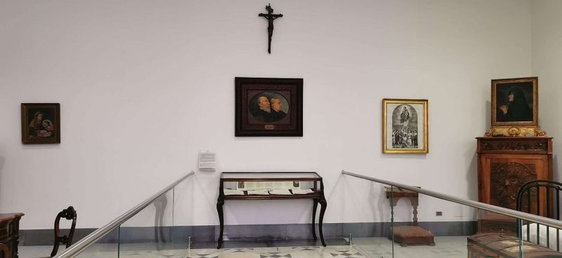 7 nuovi santi papa leone XIV da 4 continenti 4 San Bartolo Longo: Dalla Tenebra alla Luce. Ritratti storici e opere d'arte in una galleria museale di valore culturale. La parete presenta dipinti di personaggi religiosi e storici, con un crocifisso sopra, elementi tipici di un'esposizione artistica e religiosa.
