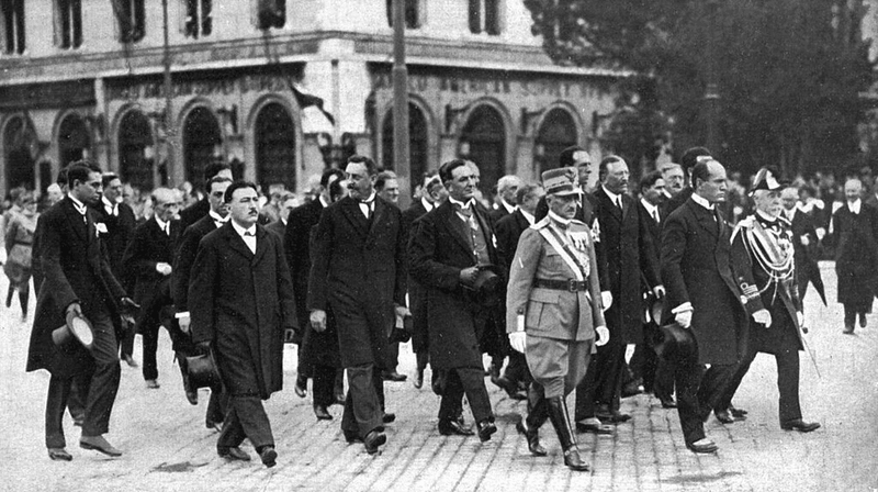 Giornata dell'Unità Nazionale e delle Forze Armate 4 novembre. 3 Uomini in abiti formali e uniformi storiche che sfilano in una piazza pubblica, scenario di un evento storico o cerimoniale, in stile vintage o d'epoca, in bianco e nero.
