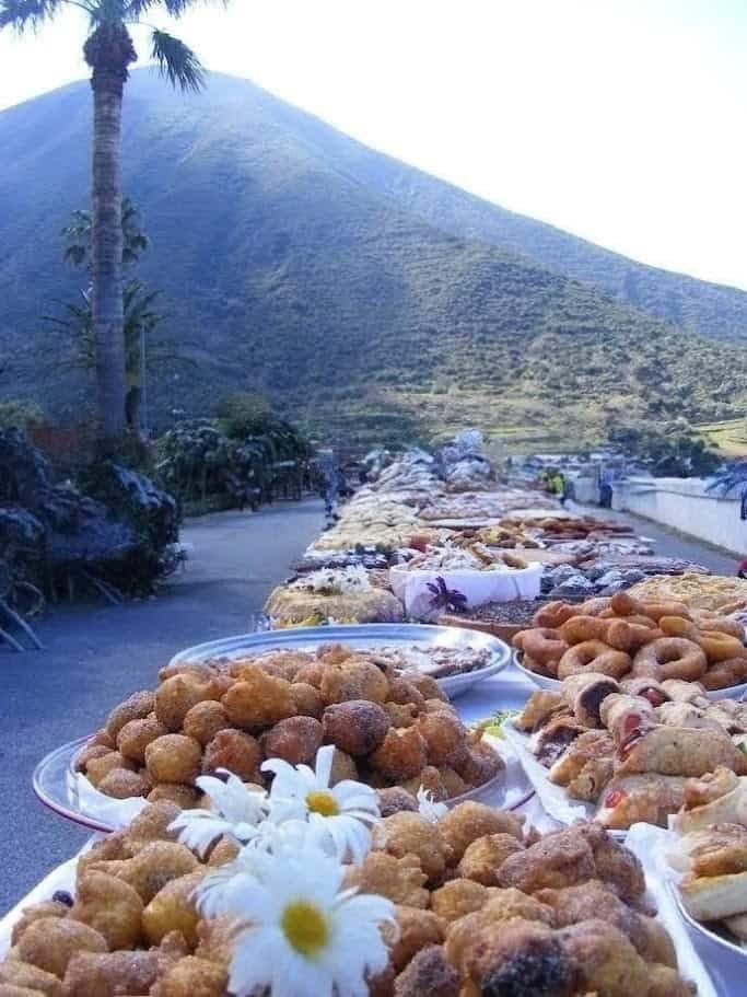 Scopri Salina isola delle Eolie, sapori autentici 5 FOTO: Banchi di street food con vari snack tradizionali messicani, con lo sfondo di un paesaggio montano e arboreo, in una scena di mercato all'aperto con montagne, palma e cielo blu.